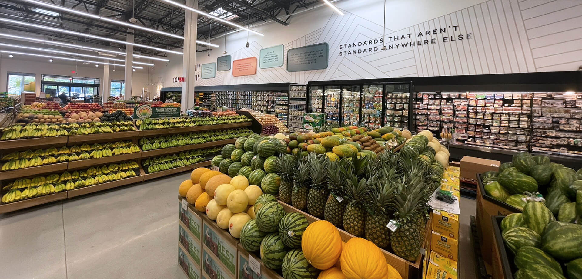 The image shows the interior of a Whole Foods Market store, specifically the produce and butcher sections. The store features signage highlighting "Quality Commitments" and a message encouraging customers to "Feel confident about what goes in your cart".