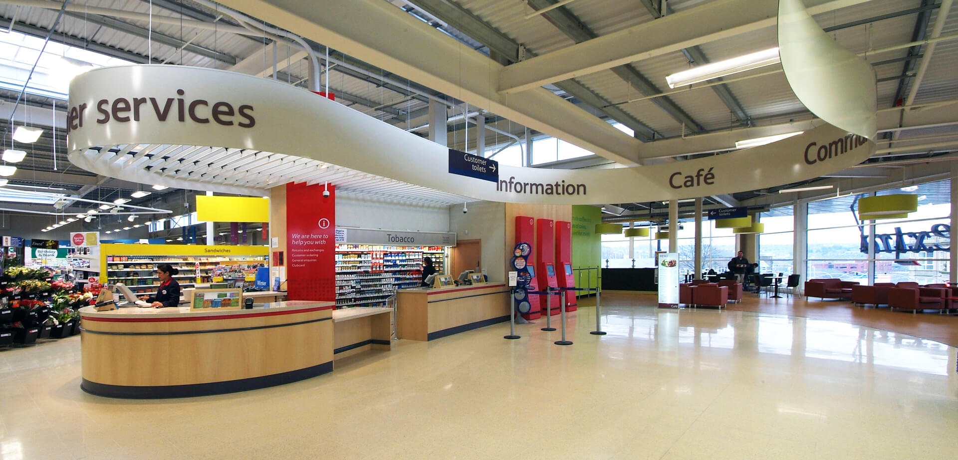 Hipermercado Tesco Extra en Accrington, Lancashire, Inglaterra. Señalización y orientación claras: Los carteles visibles y bien identificados por departamento (como los de "Atención al Cliente", "Información" y "Cafetería" en la imagen) facilitan la navegación y reducen la ansiedad de los clientes. Esto garantiza que los compradores encuentren rápidamente los artículos específicos.