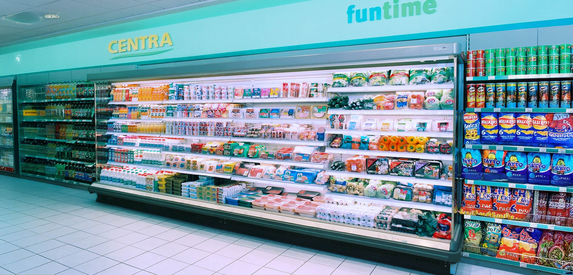 Dieses Bild zeigt den Innenraum eines Centra-Supermarkts, einer vor allem in Irland und Nordirland bekannten Einzelhandelskette. Centra ist eine große Supermarktkette mit zahlreichen Filialen in ganz Irland und bietet eine breite Palette an Lebensmitteln und Fertiggerichten. Der abgebildete Supermarkt verfügt über eine große Kühltheke mit frischem Obst und Gemüse, Fertiggerichten und Milchprodukten.