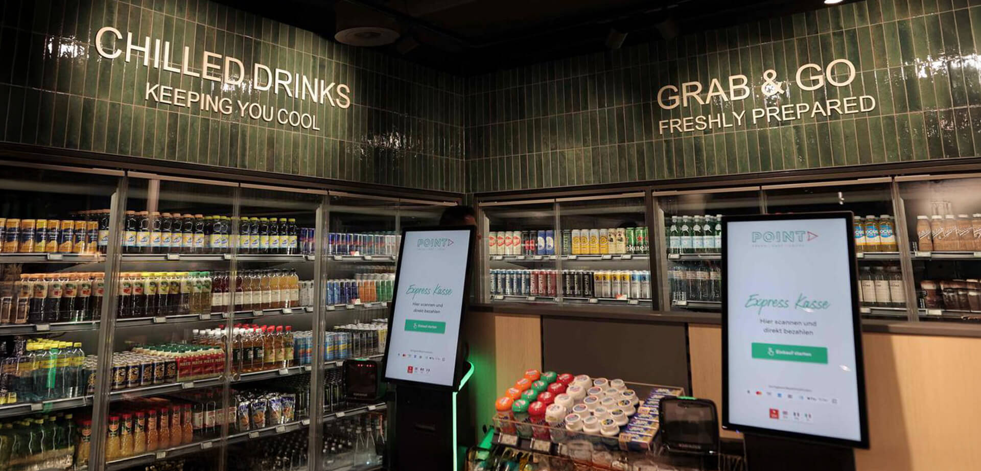 Das Bild zeigt das Innere eines SSP Point-Convenience-Stores, der speziell für Reiseanleger konzipiert ist. Der Laden setzt auf schnellen Service durch digitale Technologien wie Selbstbedienungskassen und digitale Bildschirme. Wie die Wandbeschilderung zeigt, bietet der Laden gekühlte Getränke und frisch zubereitete Speisen zum Mitnehmen an.
