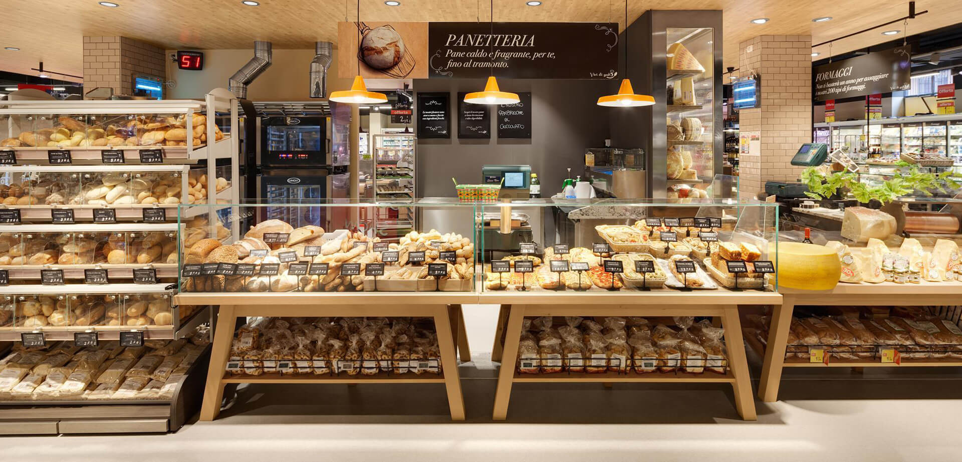 Das Bild zeigt das Innere eines Carrefour-Marktes der Feinkostabteilung in Italien, genauer gesagt die Bäckerei- und Käseabteilung. Die Schilder tragen folgende Aufschrift: PANETTERIA (Bäckerei) „Pane caldo e fragrante, per te, fino al tramonto“ (Heißes und duftendes Brot, für dich, bis Sonnenuntergang) FORMAGGI (Käse).