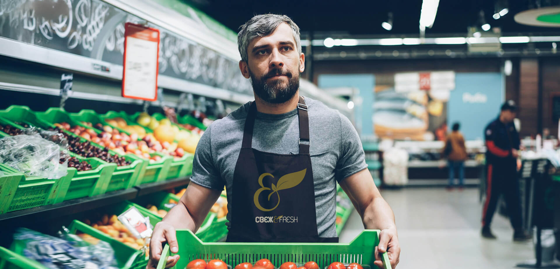 A grocery store worker holding a crate of tomatoes. The worker is wearing an apron with the logo and name "Свеж & Fresh". "Свеж & Fresh" is a brand associated with premium supermarkets.